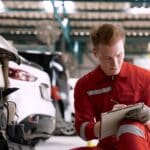 mechanic is checking front car, which enters garage in wrecked state, mechanic checks condition, writes it down in book, before taking it for repairs speed clearly know where repair.