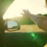 Young woman travels by car catches wind with her hand from car window. Girl with long hair is sitting in front seat of car, stretching her arm out window and catching glare of setting sun. Vacation