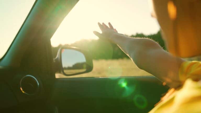 Young woman travels by car catches wind with her hand from car window. Girl with long hair is sitting in front seat of car, stretching her arm out window and catching glare of setting sun. Vacation