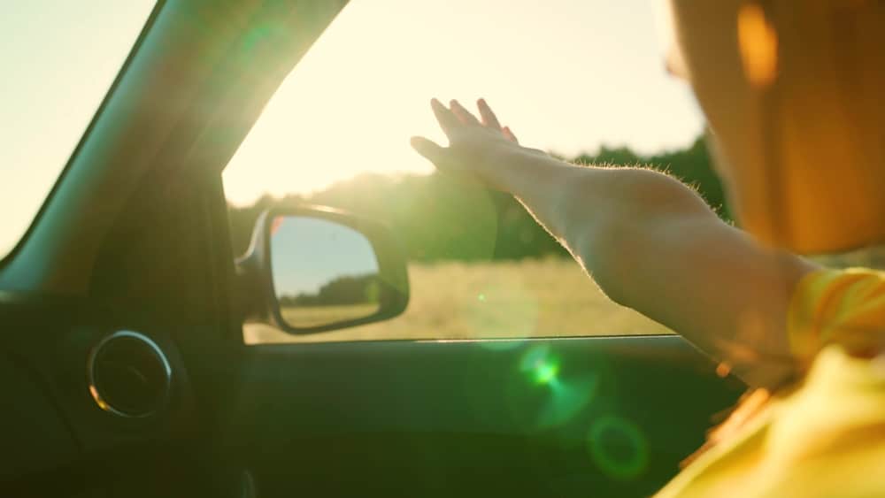 End-of-Summer Car Maintenance Tips to Prep for Fall Driving 1 Young woman travels by car catches wind with her hand from car window. Girl with long hair is sitting in front seat of car, stretching her arm out window and catching glare of setting sun. Vacation